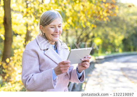 Senior woman in a sunny autumn park smiling as she uses a tablet, enjoying modern technology, relaxation and connection among golden leaves and natural light 132219897