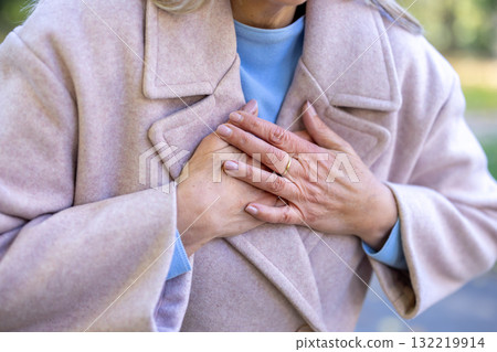 Senior woman experiencing sudden chest pain and clutching her chest with both hands, showing symptoms of a heart attack or cardiac emergency while outdoors Senior woman experiencing sudden chest pain and clutching her chest with both hands, showing symptoms of a heart attack or cardiac emergency while outdoors 132219914