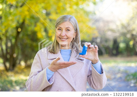 Senior woman smiling while holding a blue asthma inhaler in an autumn park, promoting respiratory health, confidence, vitality and outdoor wellbeing among older adults Senior woman smiling while holding a blue asthma inhaler in an autumn park, promoting respiratory health, confidence, vitality and outdoor wellbeing among older adults 132219918