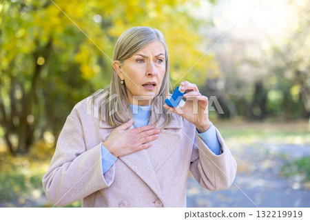 Female senior with an anxious expression experiencing an asthma attack in a park, holding an inhaler and placing a hand on her chest, seeking relief from breathing problems 132219919