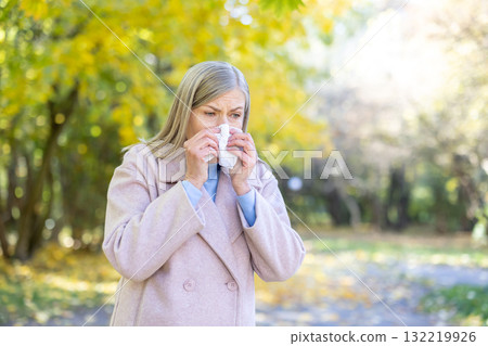 Senior woman blowing her nose into a tissue in an autumn park, battling allergy or cold symptoms amid yellow foliage, bundled in a coat and looking unwell Senior woman blowing her nose into a tissue in an autumn park, battling allergy or cold symptoms amid yellow foliage, bundled in a coat and looking unwell 132219926