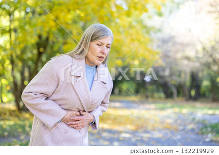 Senior woman standing in an autumn park, experiencing severe abdominal pain and stomachache, holding her stomach with both hands, focusing on discomfort 132219929