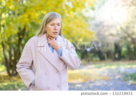 Senior woman standing in an autumn park, suffering from a sore throat and discomfort during the fall season, feeling unwell with hands touching her neck Senior woman standing in an autumn park, suffering from a sore throat and discomfort during the fall season, feeling unwell with hands touching her neck 132219931