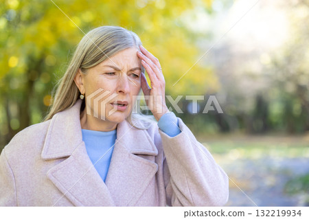 Senior woman outdoors in autumn park, pressing hand to forehead, showing migraine or headache pain, tired and stressed, pensive and unwell, upper-body portrait in daylight Senior woman outdoors in autumn park, pressing hand to forehead, showing migraine or headache pain, tired and stressed, pensive and unwell, upper-body portrait in daylight 132219934