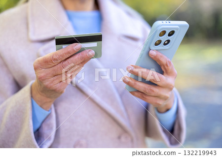 Senior woman's hands holding a credit card and a smartphone, actively engaging in cashless online shopping, banking, and digital transactions using modern technology Senior woman's hands holding a credit card and a smartphone, actively engaging in cashless online shopping, banking, and digital transactions using modern technology 132219943