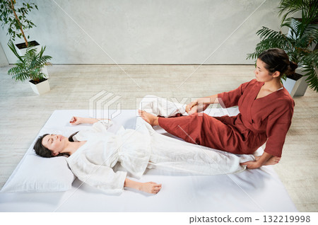 Masseuse making thai yoga massage. Woman therapist doing traditional massage treatment, stretching female patient's body. Client lying on mat, enjoying hand massage in calm atmosphere. Masseuse making thai yoga massage. Woman therapist doing traditional massage treatment, stretching female patient's body. Client lying on mat, enjoying hand massage in calm atmosphere. 132219998