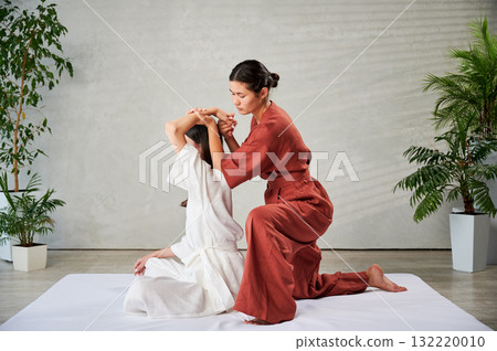 Masseuse making thai yoga massage. Woman therapist doing traditional massage treatment, stretching female patient's body. Client sitting on mat, enjoying hand massage in calm atmosphere. Masseuse making thai yoga massage. Woman therapist doing traditional massage treatment, stretching female patient's body. Client sitting on mat, enjoying hand massage in calm atmosphere. 132220010