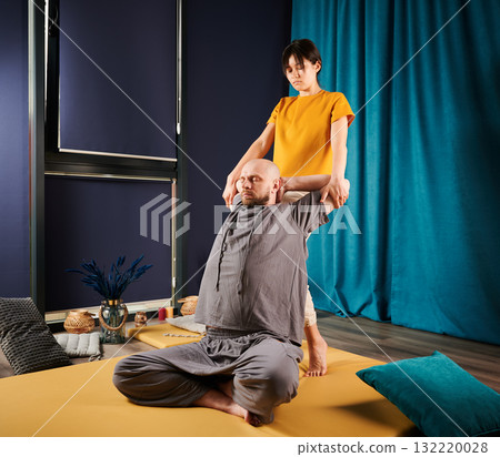 Masseuse making thai yoga massage. Woman therapist doing traditional massage treatment, stretching male patient's body. Bald client sitting on mat, enjoying hand massage in calm atmosphere. Masseuse making thai yoga massage. Woman therapist doing traditional massage treatment, stretching male patient's body. Bald client sitting on mat, enjoying hand massage in calm atmosphere. 132220028