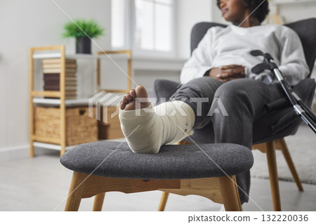 Cropped view of woman with leg in plaster bandage and crutch sittingin armchair at home Cropped view of woman with leg in plaster bandage and crutch sittingin armchair at home 132220036