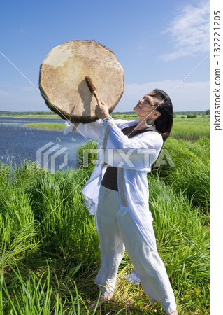 Female shaman in the white dress drumming in the natural environment Female shaman in the white dress drumming in the natural environment 132221205