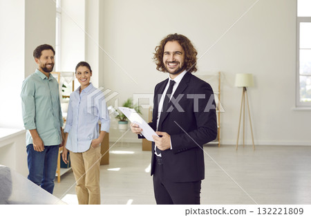 Young man realtor standing with happy couple looking at camera in new house. Young man realtor standing with happy couple looking at camera in new house. 132221809