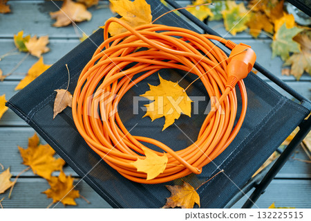 Bright orange extension cord coiled on a black mesh stool among scattered autumn leaves on a wooden deck. Outdoor tech safety, seasonal preparedness, electrical gear, autumn yard work 132225515