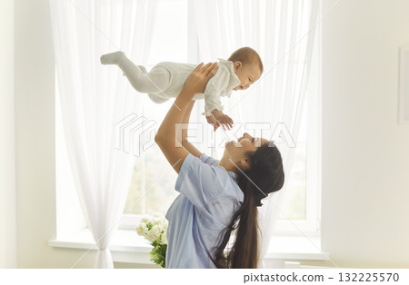 Smiling mother lifting baby by sunlit window in bright airy bedroom at home 132225570