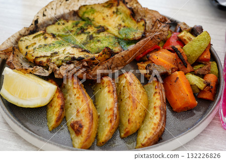 A delicious meal of baked fish with herbs, roasted potato wedges, grilled vegetables, and a lemon wedge on a plate A delicious meal of baked fish with herbs, roasted potato wedges, grilled vegetables, and a lemon wedge on a plate 132226826
