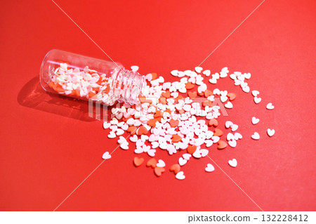 Heart-shaped sugar topping spilling from a small bottle. Cute background decoration. Overflowing feelings. Confectionery ingredients. Heart-shaped sugar topping spilling from a small bottle. Cute background decoration. Overflowing feelings. Confectionery ingredients. 132228412