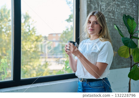 Portrait of young beautiful woman with cup looking at camera, near window Portrait of young beautiful woman with cup looking at camera, near window 132228796
