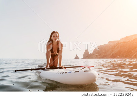 Woman Paddleboard Ocean - A woman in a black bikini kneels on a white paddleboard in the ocean, with rocky cliffs in the background. Woman Paddleboard Ocean - A woman in a black bikini kneels on a white paddleboard in the ocean, with rocky cliffs in the background. 132229242