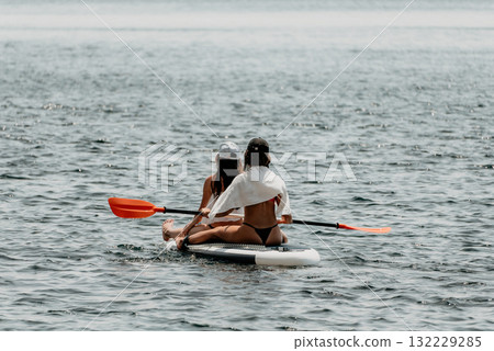 Two women floating on a surfboard in the ocean. One of them is wearing a white shirt and the other is wearing a black bikini. Two women floating on a surfboard in the ocean. One of them is wearing a white shirt and the other is wearing a black bikini. 132229285