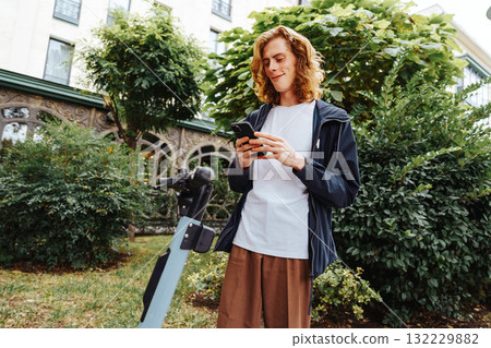 Young man checking phone while standing near electric scooter in green park area 132229882
