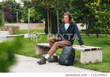 Young man enjoys music on a park bench in a lush green outdoor space during daytime 132229922