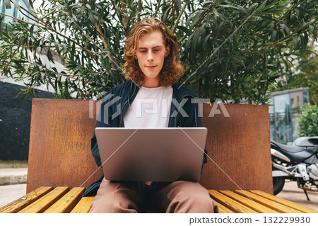 Young man works on laptop while sitting on a bench under green leaves in an urban setting Young man works on laptop while sitting on a bench under green leaves in an urban setting 132229930