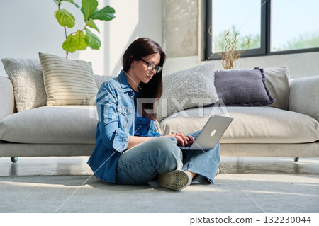 Young serious woman typing on laptop sitting on floor near couch at home Young serious woman typing on laptop sitting on floor near couch at home 132230044