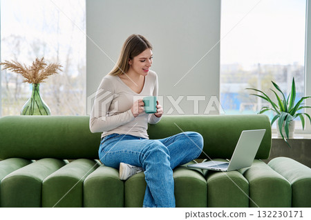 Young relaxing woman with cup looking at laptop screen, sitting on sofa at home Young relaxing woman with cup looking at laptop screen, sitting on sofa at home 132230171