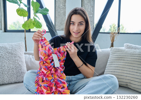 Young woman unpacking cardboard box parcel with online order, looking at new clothes 132230260