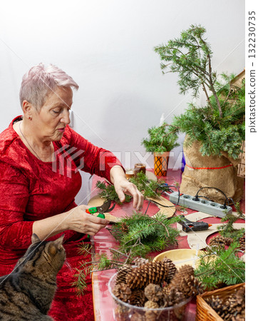Senior old woman and cat craft. Grandmother make Christmas wreath from pine branches. Home decoration in December. Winter Holiday preparing 132230735