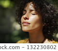 Woman inhaling deeply in nature, curly hair bright. Calm image captures wellness and tranquility in green woods. 132230802