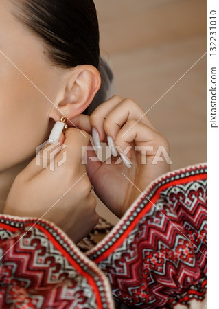 Woman Putting On Gold Earrings, Close-Up, Red Embroidered Top, Delicate Jewelry, Fashion Accessory, Stylish Lifestyle, Warm Tones, Intricate Detail. 132231010