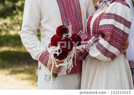 Ukrainian Wedding Ceremony: Traditional Embroidered Outfits, Red Dahlia Bouquet, Couple Embracing, Natural Outdoor Setting, Rustic Style. Ukrainian Wedding Ceremony: Traditional Embroidered Outfits, Red Dahlia Bouquet, Couple Embracing, Natural Outdoor Setting, Rustic Style. 132231015