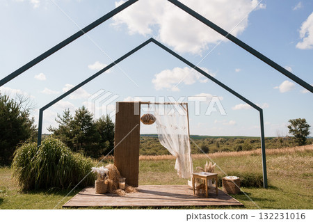 Outdoor Wedding Ceremony Setup with Rustic Arch, Natural Backdrop, and Bohemian Decor in a Grassy Field Under a Blue Sky. 132231016