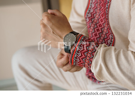 Man Adjusting Minimalist Black Watch with Leather Strap Wearing Traditional Embroidered Vest, Neutral Tones, Stylish Accessory, Close-Up Shot. Man Adjusting Minimalist Black Watch with Leather Strap Wearing Traditional Embroidered Vest, Neutral Tones, Stylish Accessory, Close-Up Shot. 132231034