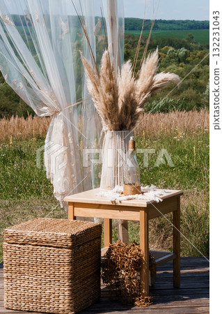 Bohemian Wedding Decor Scene with Pampas Grass, Rustic Wooden Furniture, Sheer Curtains and Summer Field Backdrop Natural Texture and Warm Tones Bohemian Wedding Decor Scene with Pampas Grass, Rustic Wooden Furniture, Sheer Curtains and Summer Field Backdrop Natural Texture and Warm Tones 132231043