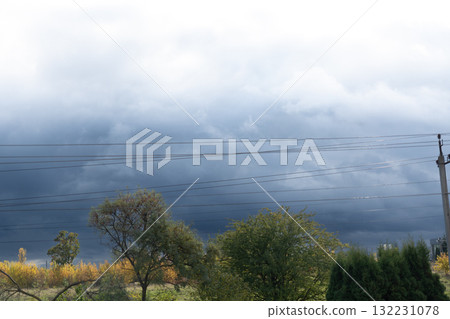 Dramatic Storm Clouds Over Trees and Power Lines Dark Blue Sky, Autumn Foliage, Rural Landscape, Ominous Weather, Atmospheric Perspective. Dramatic Storm Clouds Over Trees and Power Lines Dark Blue Sky, Autumn Foliage, Rural Landscape, Ominous Weather, Atmospheric Perspective. 132231078