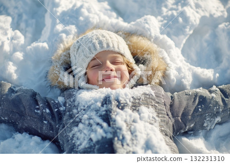 A joyful child lies in soft, fresh snow with arms spread wide, creating snow angels under clear blue skies. The winter scene showcases bright sunlight reflecting off the snow 132231310