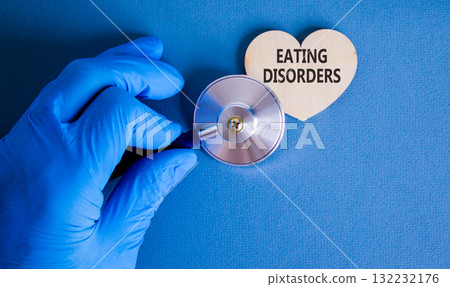 Eating disorders symbol. Concept words Eating disorders on beautiful wooden heart. Doctor's hand in blue medical glove. Beautiful blue background. Medical healthy lifestyle eating disorders concept. Eating disorders symbol. Concept words Eating disorders on beautiful wooden heart. Doctor's hand in blue medical glove. Beautiful blue background. Medical healthy lifestyle eating disorders concept. 132232176