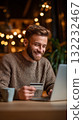 Young Man Smiles While Shopping Online in a Cozy Cafe During Evening Hours 132232467