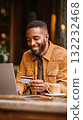 Young Man Smiles While Shopping Online in a Cozy Cafe During Evening Hours 132232468