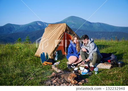 Scenic mountain campsite with two tents in morning. Two campers enjoying serene view of rolling mountains under clear sky. Picturesque sunrise landscape, creating tranquil and cozy outdoor experience. Scenic mountain campsite with two tents in morning. Two campers enjoying serene view of rolling mountains under clear sky. Picturesque sunrise landscape, creating tranquil and cozy outdoor experience. 132232948