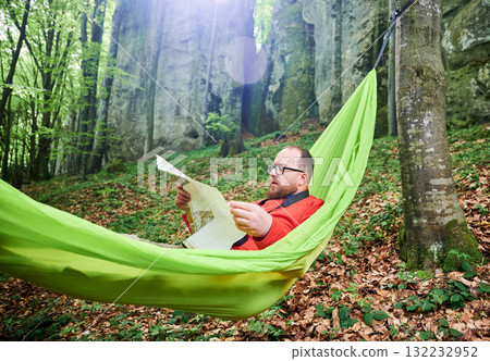 Man traveler in glasses relaxes in green hammock in lush forest. Male tourist examining map intently, surrounded by green foliage and tall rocks, planning outdoor adventure, hiking or climbing route. Man traveler in glasses relaxes in green hammock in lush forest. Male tourist examining map intently, surrounded by green foliage and tall rocks, planning outdoor adventure, hiking or climbing route. 132232952