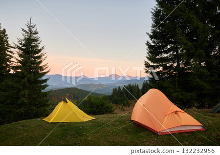Orange tent and yellow tent on grassy hillside, surrounded by tall pine trees. Scene overlooks stunning mountain range under pastel-colored sky at sunrise, creating serene, picturesque camping spot. Orange tent and yellow tent on grassy hillside, surrounded by tall pine trees. Scene overlooks stunning mountain range under pastel-colored sky at sunrise, creating serene, picturesque camping spot. 132232956