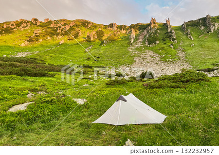 Lone white tourist tent in lush green valley surrounded by towering hills and rocky cliffs. Sky above shows hint of sunrise colors, creating serene camping scene. Carpathian mountains, Ukraine. 132232957