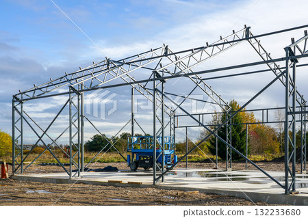 Steel frame of industrial warehouse under construction with scaffolding and blue lift on a sunny day 132233680