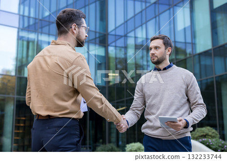 Two professional businessmen shaking hands in an urban setting, solidifying their partnership and reaching a positive agreement outside a modern office building 132233744