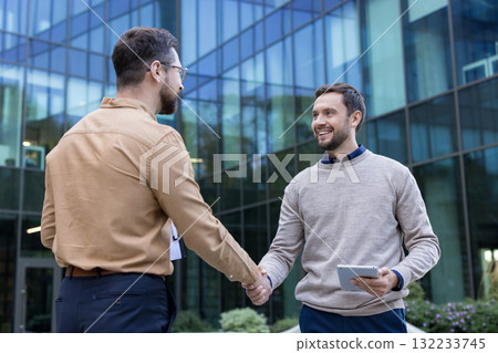 Two smiling men in smart casual attire meeting and shaking hands outside a contemporary glass-fronted office building, completing a successful agreement Two smiling men in smart casual attire meeting and shaking hands outside a contemporary glass-fronted office building, completing a successful agreement 132233745