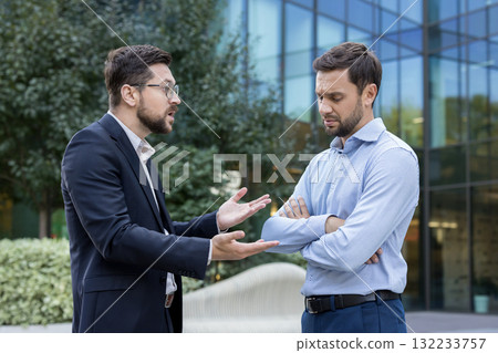 Two businessmen argue heatedly outside a modern office building, one gesturing angrily while the other listens with frustration, capturing workplace conflict and tension Two businessmen argue heatedly outside a modern office building, one gesturing angrily while the other listens with frustration, capturing workplace conflict and tension 132233757