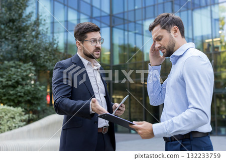 Two businessmen having an intense argument and conflict outdoors, one appearing stressed and covering his head while holding documents, the other gesturing as he speaks Two businessmen having an intense argument and conflict outdoors, one appearing stressed and covering his head while holding documents, the other gesturing as he speaks 132233759