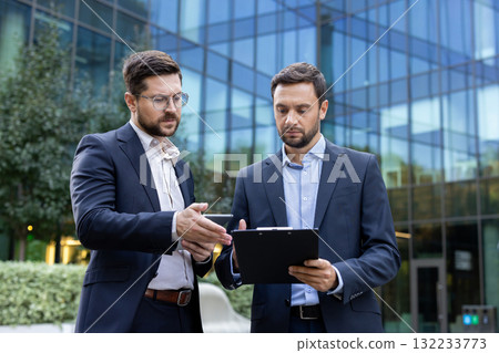 Two professional businessmen are collaborating and planning a new project, looking at a tablet and clipboard while standing in front of a contemporary glass office building Two professional businessmen are collaborating and planning a new project, looking at a tablet and clipboard while standing in front of a contemporary glass office building 132233773